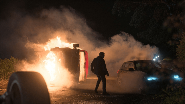An undisclosed person surrounded by crashed, flaming and smoking cars at night, in a still from Corriedale.